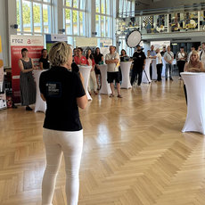Großer Messe- oder Veranstaltungssaal mit vielen Besuchern, die sich in Gruppen unterhalten. Zwei Personen im Vordergrund präsentieren oder führen durch den Raum. Im Hintergrund stehen Infostände mit Werbematerialien und Logos.