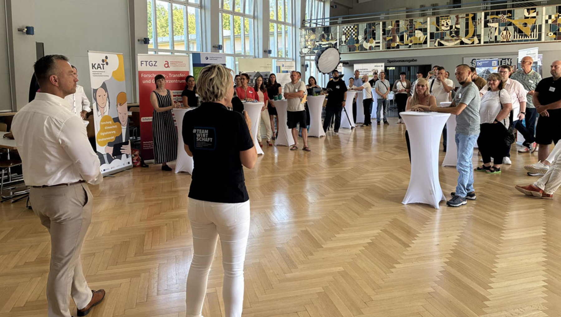 Großer Messe- oder Veranstaltungssaal mit vielen Besuchern, die sich in Gruppen unterhalten. Zwei Personen im Vordergrund präsentieren oder führen durch den Raum. Im Hintergrund stehen Infostände mit Werbematerialien und Logos.