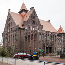 Historisches Backsteingebäude mit steilem Satteldach, Zwerchhäusern und Erkern, umgeben von einem schmiedeeisernen Zaun. Vor dem Eingang steht ein roter Kleinwagen auf der Straße.