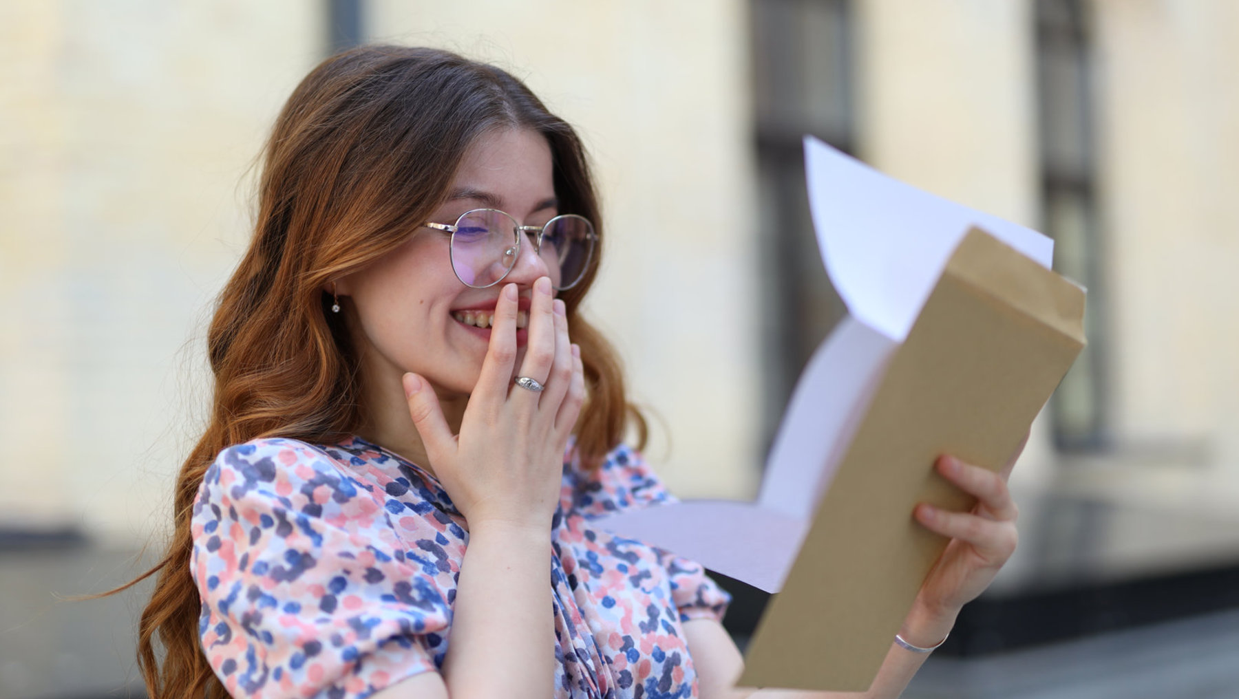 Eine Person mit Brille und langem Haar hält überrascht ein braunes Kuvert mit einem weißen Umschlag in der Hand und lacht glücklich.