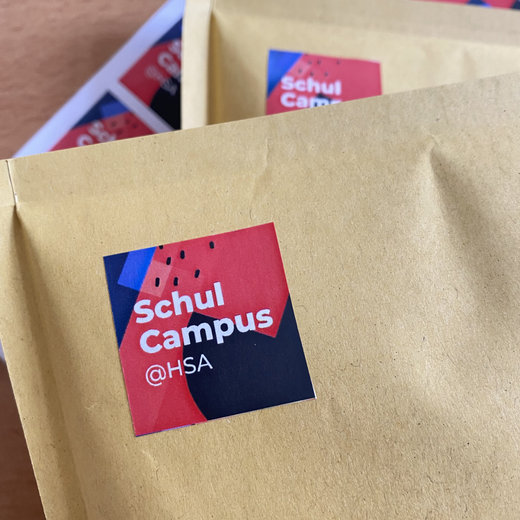 Brauner Umschlag mit Aufkleber des „Schul Campus“ der HSA, bestehend aus rotem und blauem Feld mit weißer Schrift.