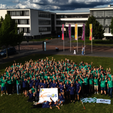 Große Gruppe von Menschen in grünen T-Shirts, die auf einem Rasen vor einem Gebäudekomplex posiert, einige halten ein Schild mit dem Logo „Copernicus“. Drei Flaggen im Hintergrund.