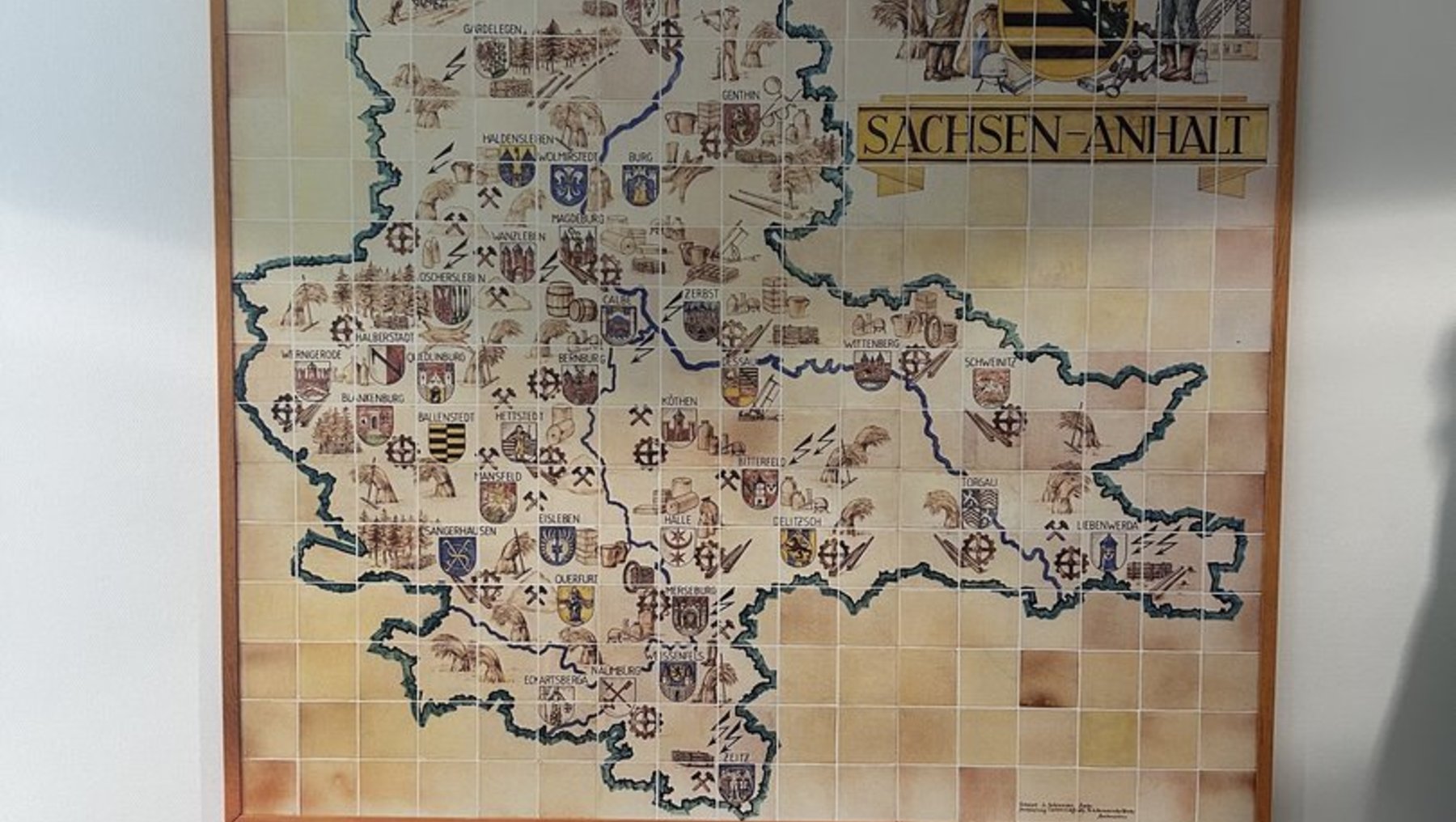 Karte des Bundeslandes Sachsen-Anhalt mit den Umrissen der Landkreise und Städte, die mit historischen Wappen und Symbolen verziert sind. Oben rechts abgebildet sind zwei Personen mit einem Fachwerkhaus im Hintergrund.