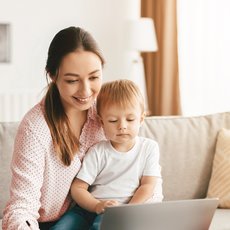 Eine lächelnde Frau sitzt mit einem kleinen Kind auf dem Schoß auf einem Sofa und zeigt ihm gemeinsam ein Laptopbildschirm. Beide wirken konzentriert und glücklich.