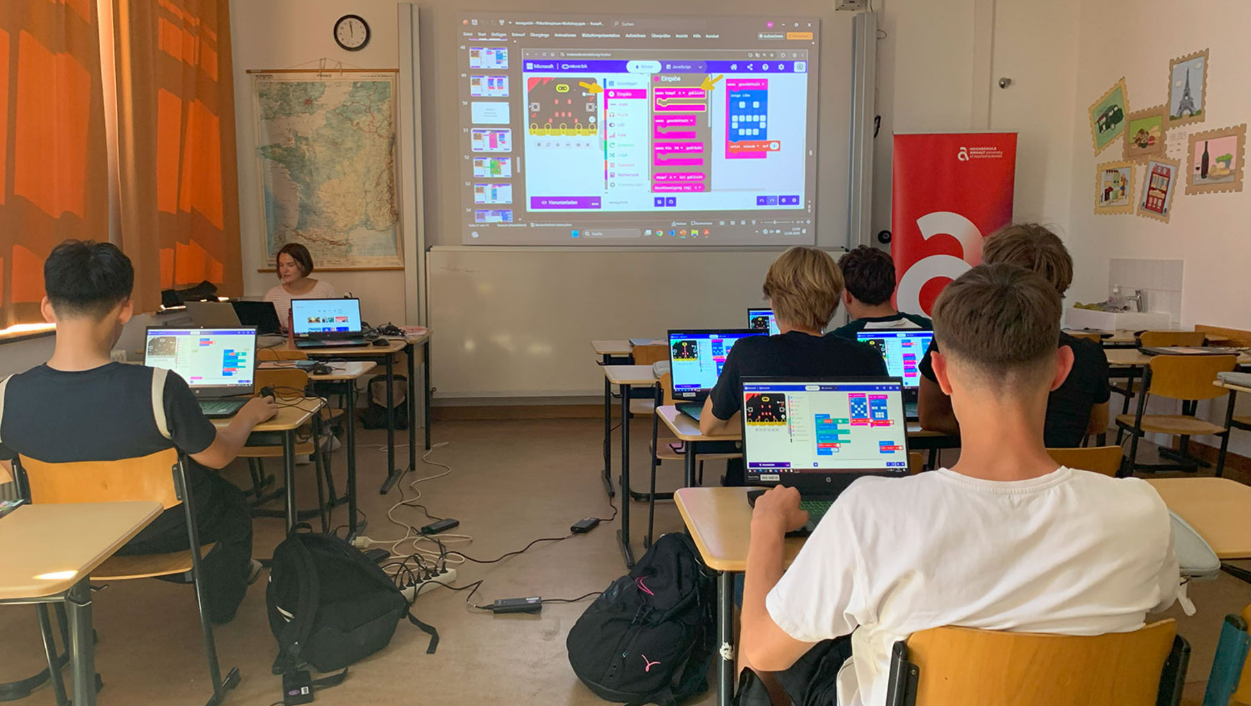 Ein Klassenzimmer mit Schülern, die an Laptops sitzen und gemeinsam ein Programm mit farbigen Code-Blöcken auf einem großen Bildschirm am Ende des Raumes bearbeiten.