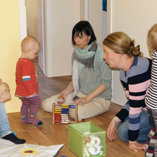 A group of adults and children are gathered in a room, engaging in play and conversation. They are sitting on the floor, surrounded by toys and educational materials. The atmosphere appears to be warm and interactive, with both adults and children participating in the activities.