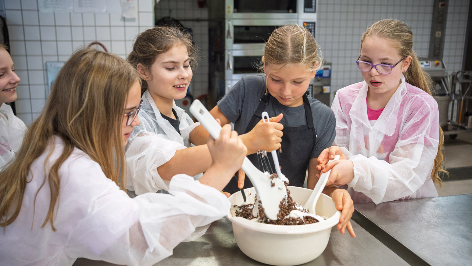 Vier Mädchen in weißer Kochkleidung stehen um eine große Schüssel mit Teig und mixen gemeinsam mit großen Holzlöffeln, während sie sich konzentriert unterhalten. Eine Person im Hintergrund beobachtet.