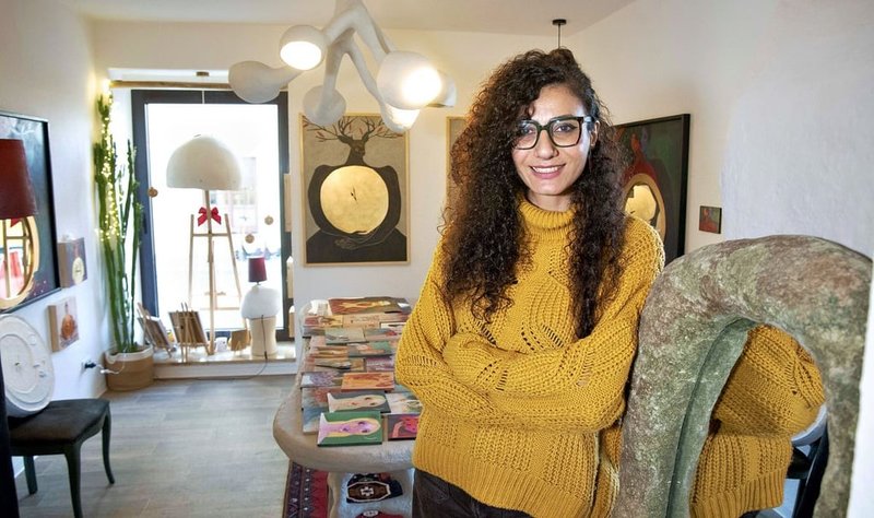 Eine lächelnde Person mit lockigen Haaren, Brille und gelbem Pullover sitzt entspannt auf einem Sessel in einem gemütlichen Raum mit Büchern und Deko im Hintergrund.