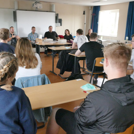 Ein Seminarraum mit etwa 15 Teilnehmenden, die an Tischen sitzen und einer Diskussion folgen. Im Vordergrund sitzen mehrere Personen mit Blick zur Gruppe.