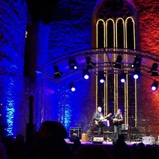 A live concert in a large, dimly lit venue with colorful lighting. Two musicians are performing on stage with a grand pipe organ behind them. The audience is watching attentively from the foreground. The atmosphere is vibrant and engaging.