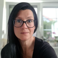 Eine Person mit dunklem, mittellangem Haar und Brille, die freundlich in die Kamera schaut, trägt ein schwarzes Oberteil und wirkt entspannt.