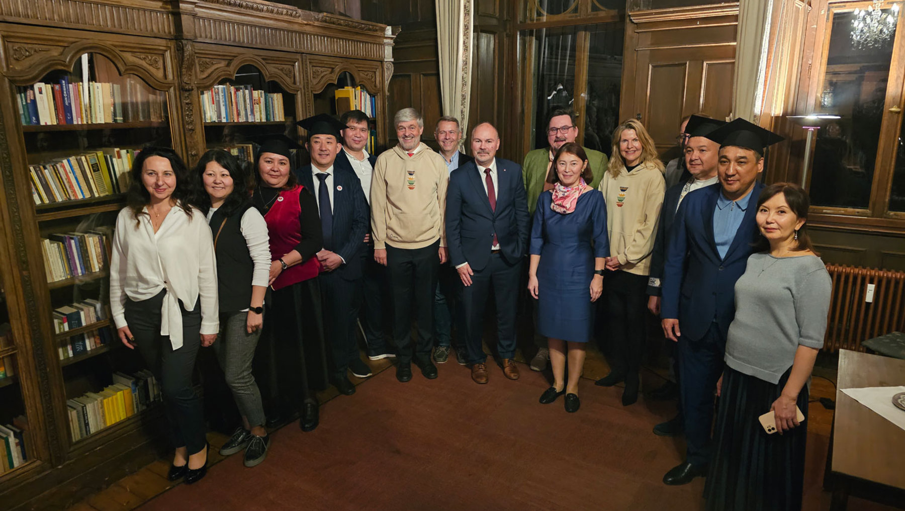 Eine Gruppe von Menschen steht in einem Raum mit Bücherregalen und Holzvertäfelungen. Sie lächeln in die Kamera und tragen formelle Kleidung. Die Atmosphäre wirkt feierlich und freundlich.