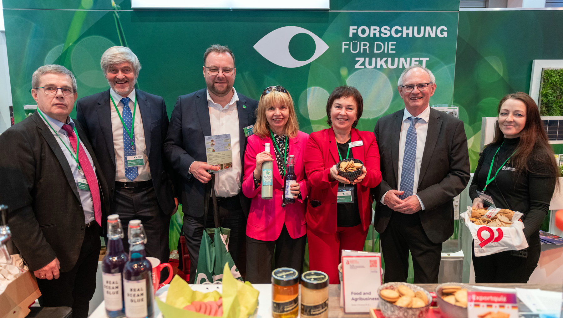 Sechs Personen stehen lächelnd vor einem Infostand mit dem Schriftzug „Forschung für die Zukunft“ und halten regionale Produkte wie Bier, Brot und Marmelade. Im Hintergrund sind Logos von Otto-von-Guericke-Universität Magdeburg und Hochschule Harz zu sehen.