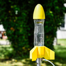 Gelber Vogeltränker in Form einer Rakete mit durchsichtigem Wasserbehälter und gelber Spitze, montiert auf einem Metallstab im Garten.