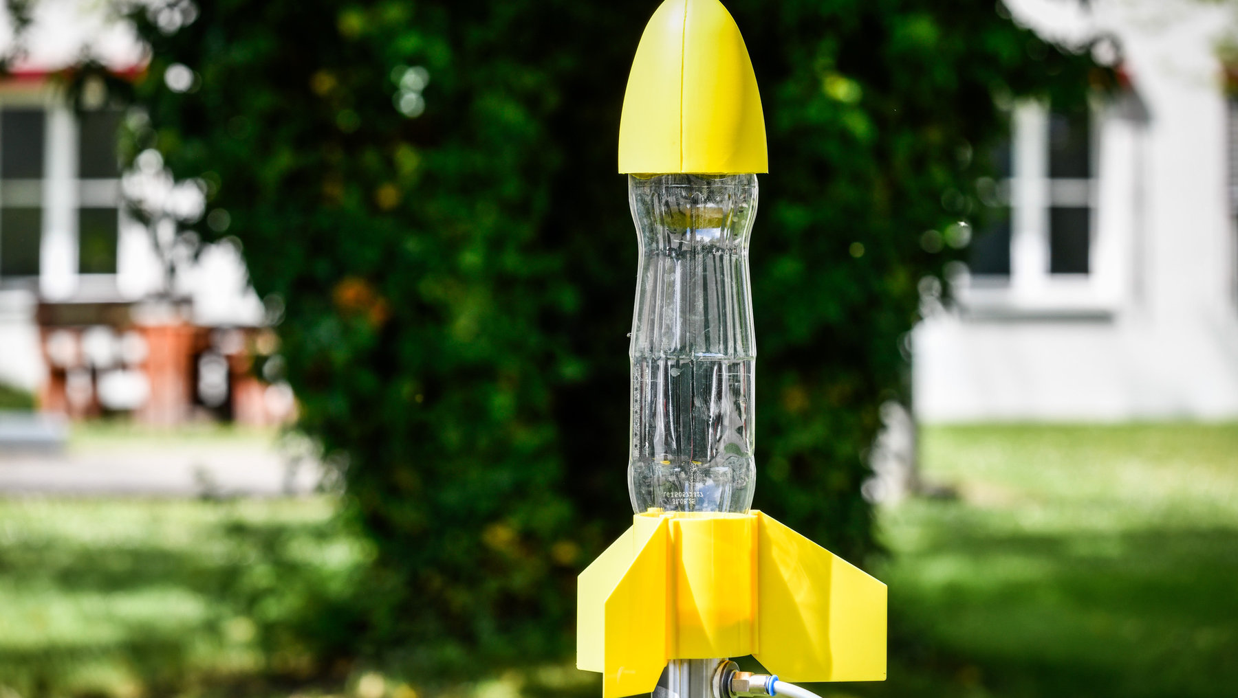 Gelber Vogeltränker in Form einer Rakete mit durchsichtigem Wasserbehälter und gelber Spitze, montiert auf einem Metallstab im Garten.