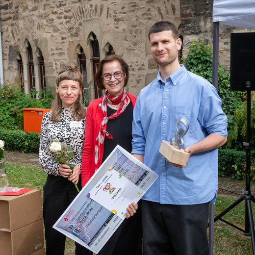 Drei Personen vor historischer Mauer: Eine Frau hält eine Urkunde mit Logo „TAG der LEHRE 2026“, eine zweite Frau hält eine weiße Blume, ein Mann hält eine Trophäe in Form eines Buches. Alle lächeln und wirken zufrieden.