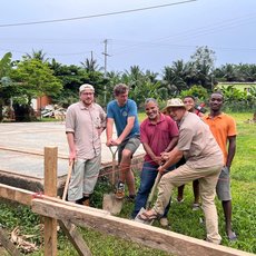 Fünf Männer arbeiten gemeinsam an der Errichtung eines Zauns oder einer Holzkonstruktion in ländlicher Umgebung, indem sie einen Pfosten mit Seilen und Hebelwirkung in den Boden rammen.