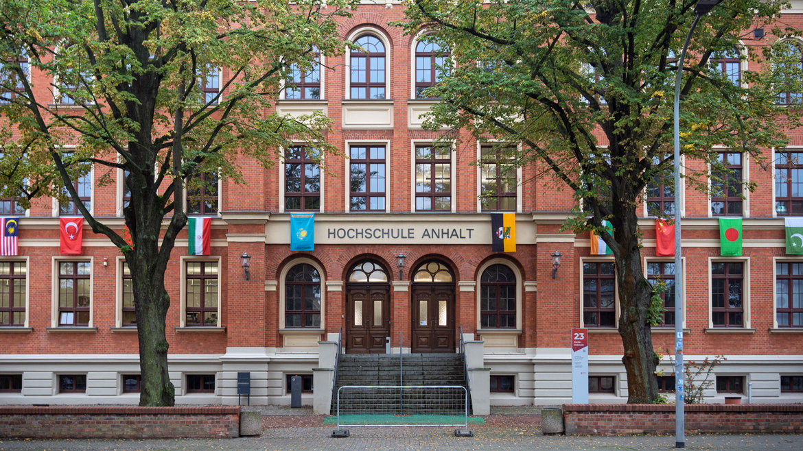 Rotes Backsteingebäude mit zentralem Haupteingang und Treppe, beschriftet als „Hochschule Anhalt“. Mehrere Flaggen internationaler Partnerhochschulen hängen an der Fassade.