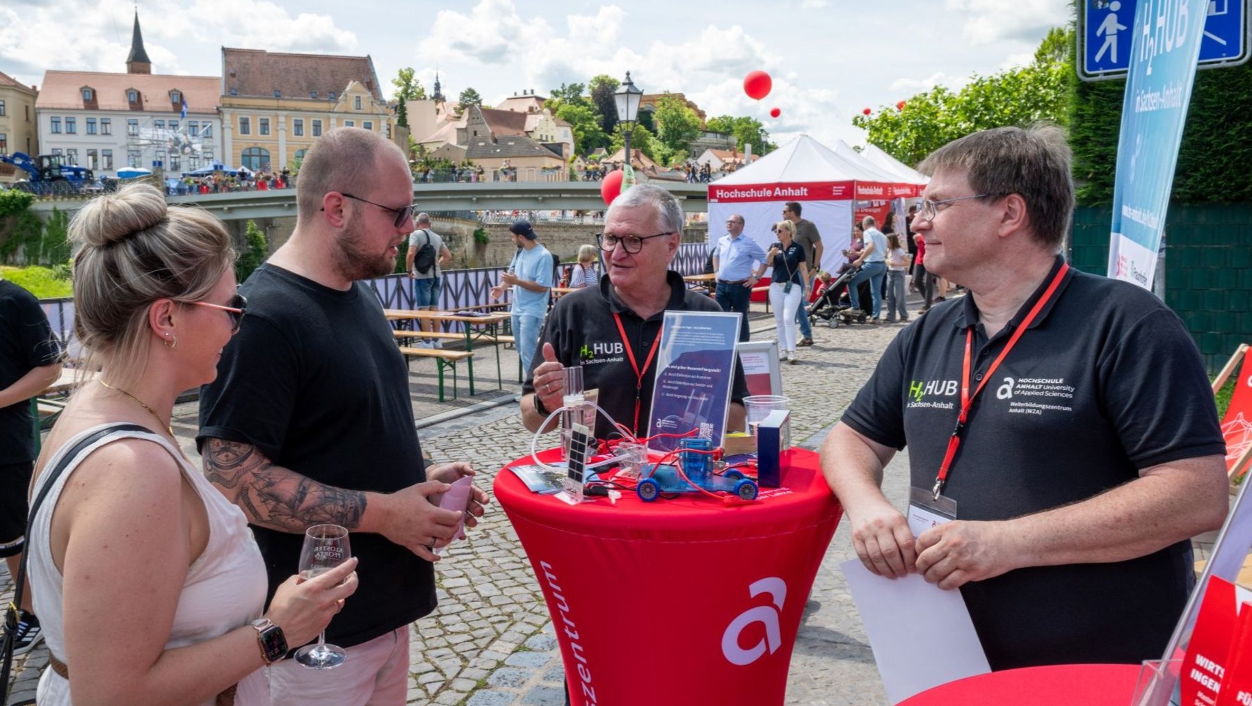 Drei Personen stehen an einem roten Infostand mit dem Logo der Hochschule Anhalt. Eine Frau in weißem Kleid und zwei Männer in schwarzen Shirts unterhalten sich, während einer der Männer Informationsmaterialien aushändigt. Im Hintergrund sind weitere Besucher und ein Zelt zu sehen.