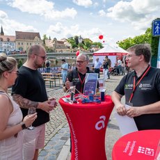 Drei Personen stehen an einem roten Infostand mit dem Logo der Hochschule Anhalt. Eine Frau in weißem Kleid und zwei Männer in schwarzen Shirts unterhalten sich, während einer der Männer Informationsmaterialien aushändigt. Im Hintergrund sind weitere Besucher und ein Zelt zu sehen.