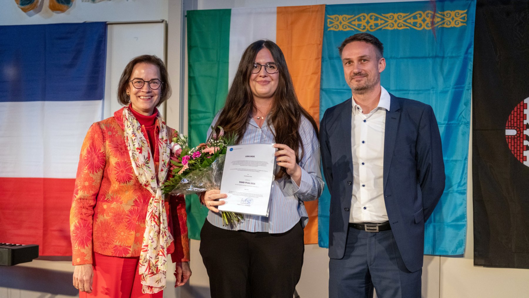 Eine Frau in der Mitte hält ein Zertifikat und einen Blumenstrauß, während sie lächelnd zwischen zwei Personen steht. Die linke Person trägt eine bunte Jacke mit Schal, die rechte einen dunklen Blazer. Im Hintergrund hängen Flaggen und ein Banner.