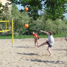 A group of five people playing beach volleyball on a sandy court. One person is jumping to hit the ball over the net, while others are positioned around the court, ready to react. The scene is set outdoors with trees and buildings in the background.