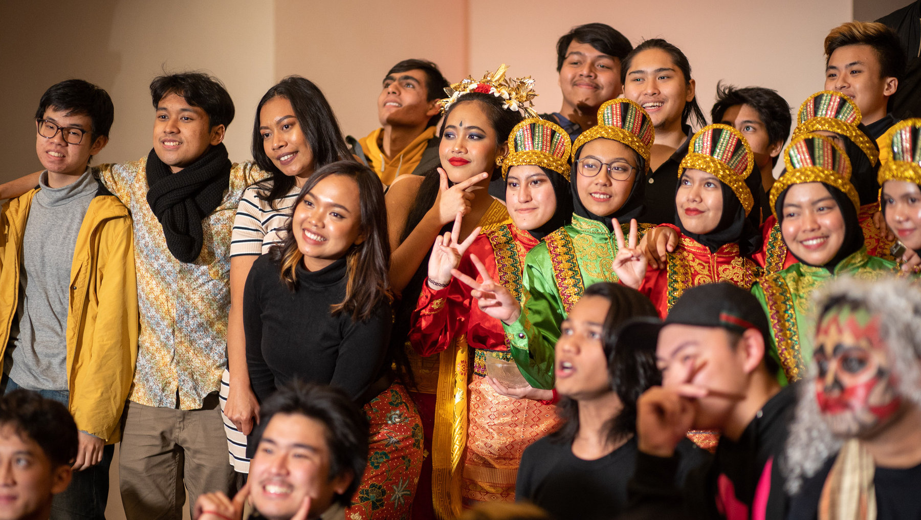 Gruppe junger Menschen, fröhlich und lässig posierend. Einige tragen traditionelle Kopfbedeckungen und bunte, festliche Kleidung, andere moderne Outfits. Viele zeigen Peace-Zeichen oder lächeln in die Kamera.