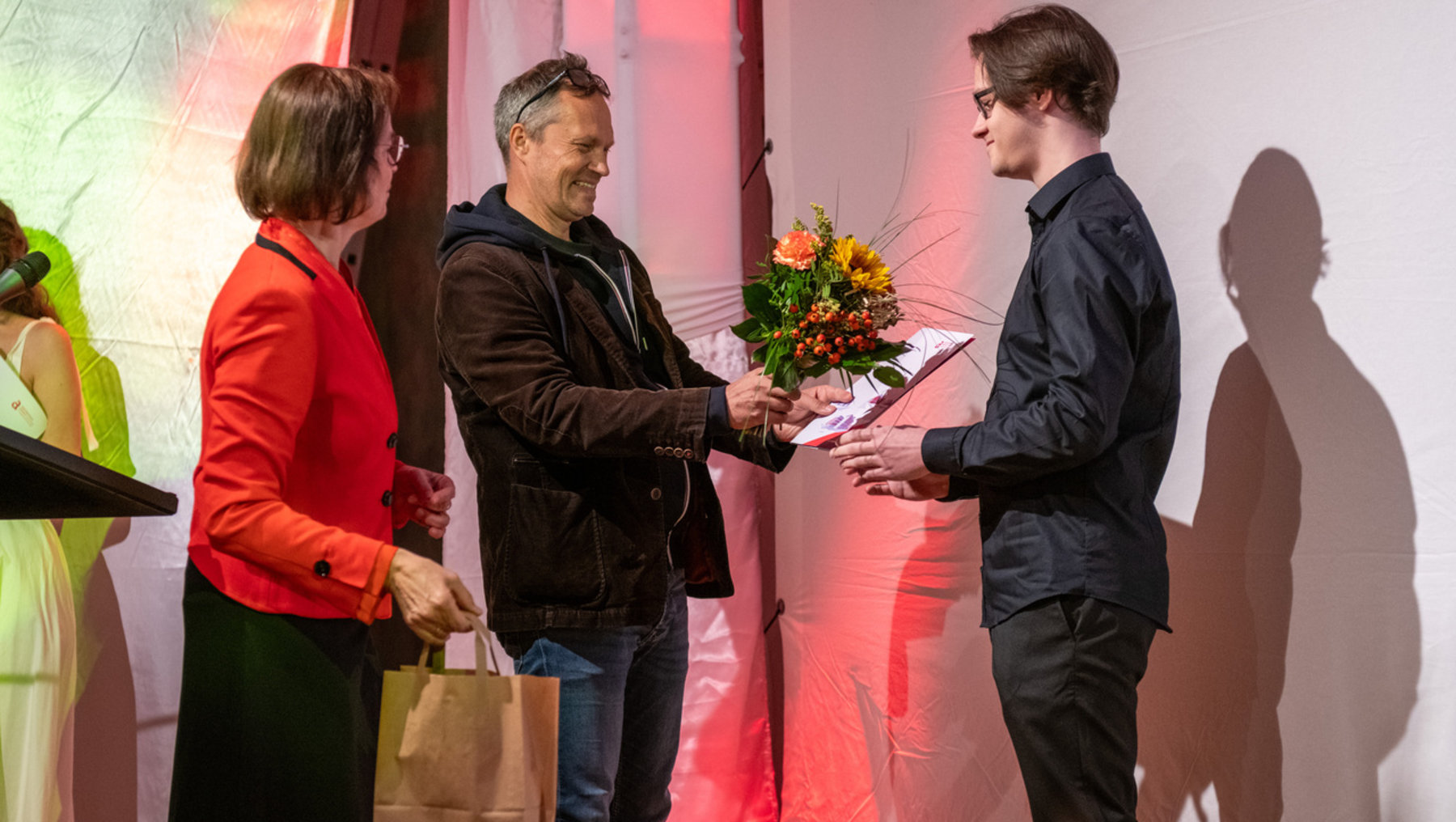 Ein Mann erhält von einer Frau einen Blumenstrauß und eine Urkunde überreicht, während ein weiterer Mann danebensteht und eine Tüte hält. Die Szene wirkt feierlich und ehrend.
