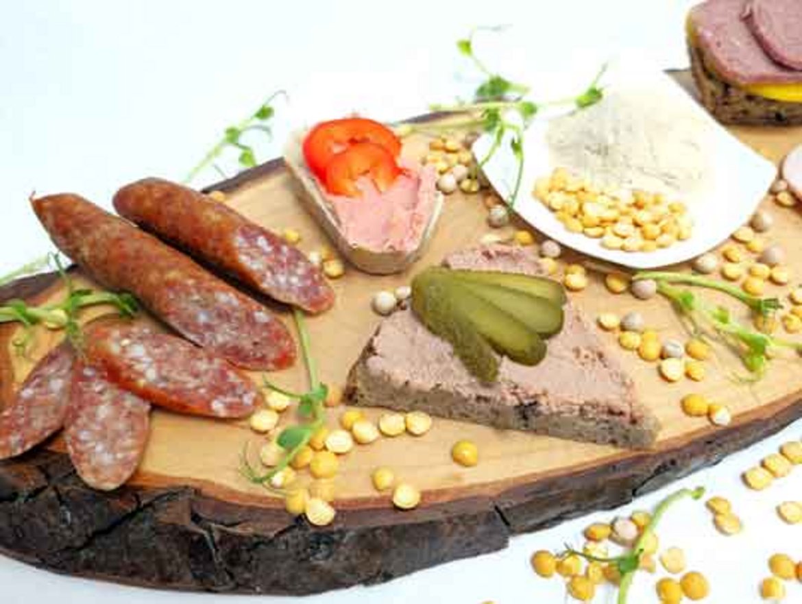 Drei würstchenartige salami-scheiben, eine scheibe roher schinken und eine scheibe gepökelter fleischspeck auf einem brett. Dazu eine kleine schüssel mit gelben maiskörnern, ein tomatenstück und ein paar frische kräuterblätter.