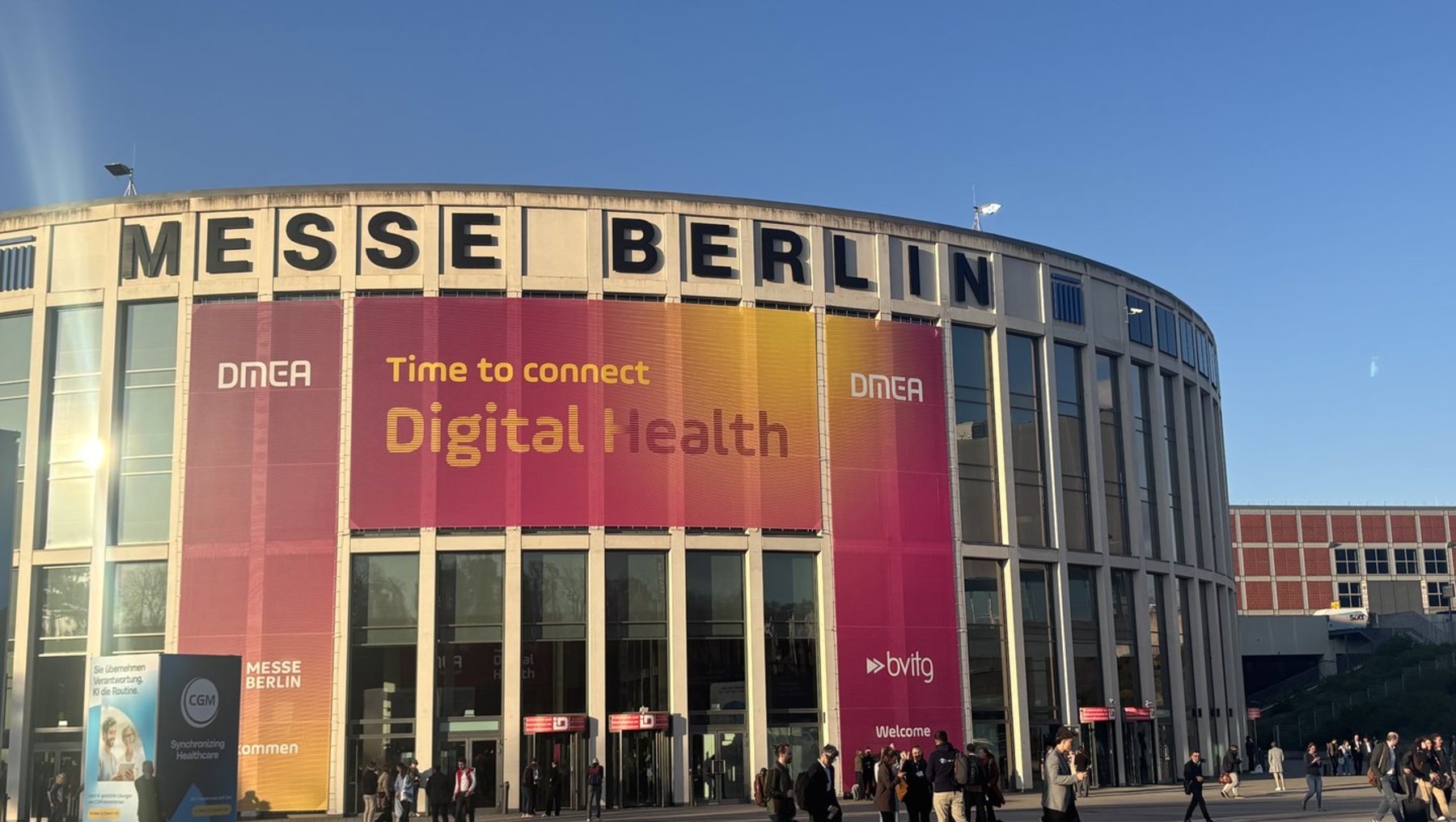 Großes Messegebäude mit der Aufschrift „Messe Berlin“. Vor dem Eingang ein Banner mit der Überschrift „Time to connect Digital Health“ und dem Logo DMEA. Menschen betreten oder verlassen das Gebäude.