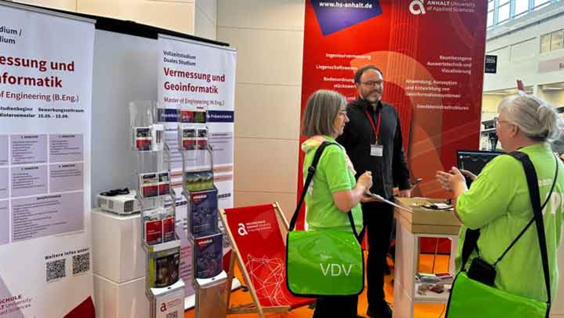 Drei Personen stehen an einem Messestand zu den Themen Vermessung und Geoinformatik. Eine Person mit grünem VDV-Shirt erklärt Inhalte, während zwei andere zuhören. Im Hintergrund sind Informationsplakate und ein Laptop zu sehen.