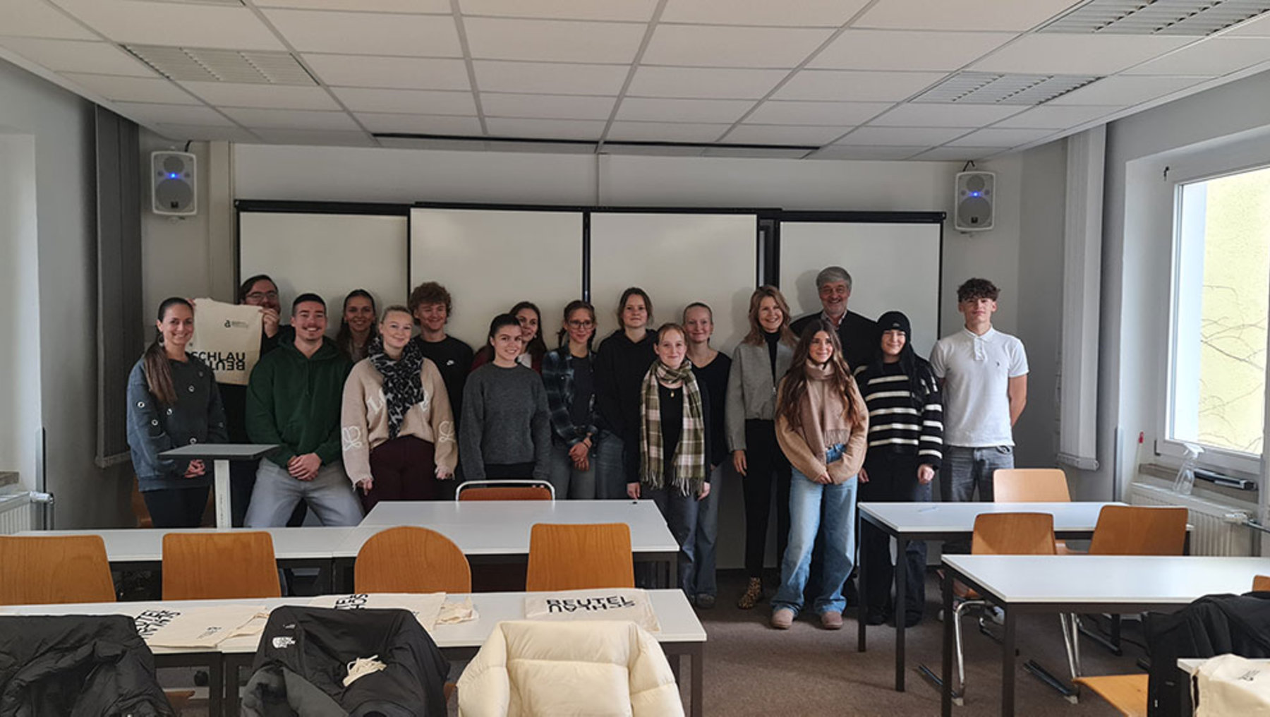 Gruppe von 16 Personen, darunter Männer und Frauen, steht lächelnd in einem hellen Seminarraum mit Tischen, Stühlen und Whiteboard. Einige halten ein Plakat mit Aufschrift, andere stehen locker daneben.