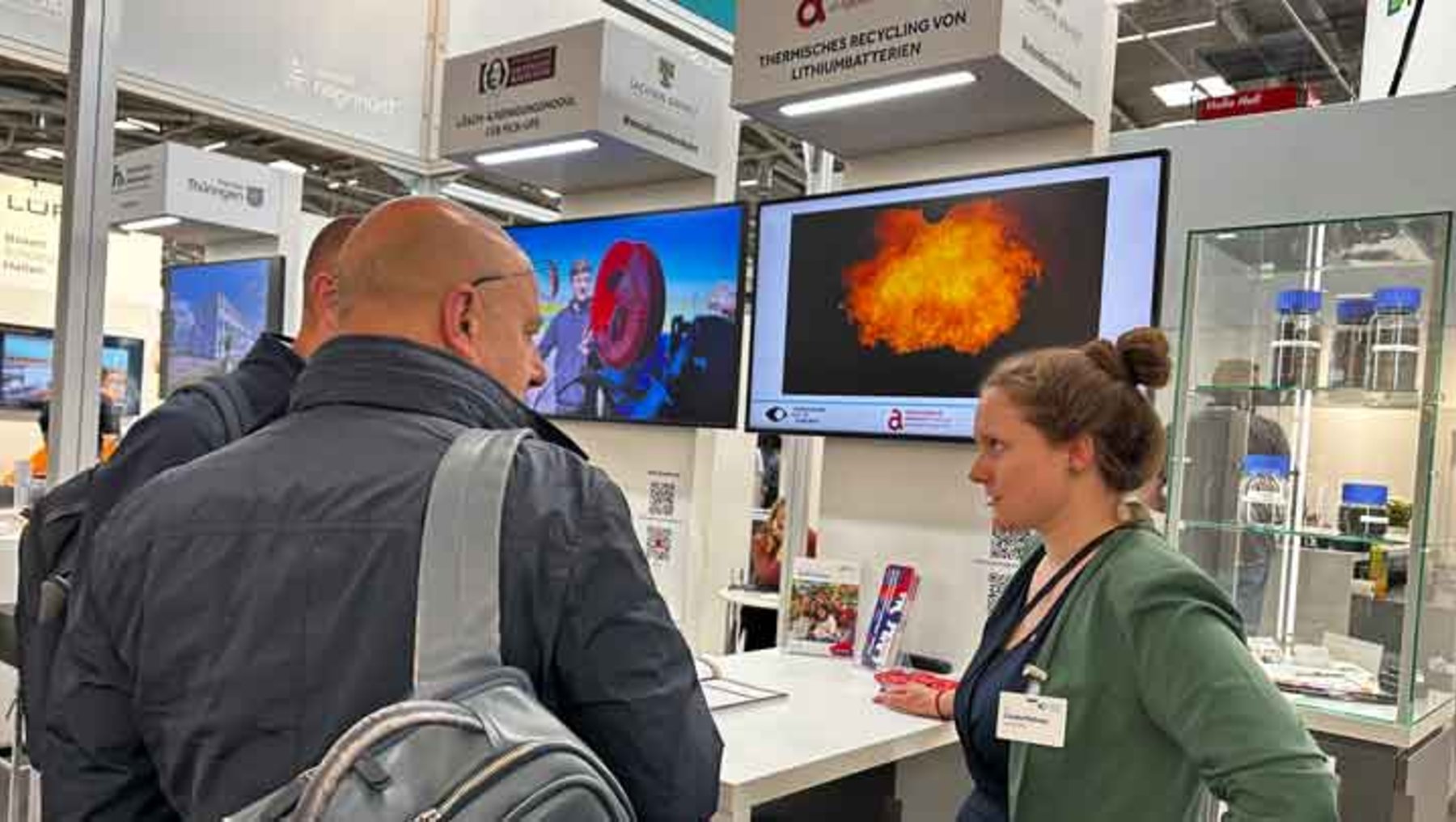 Zwei Personen auf einer Messe: Ein Mann mit Rucksack betrachtet mit einer Frau in grünem Shirt und Namensschild eine große Anzeige mit einer Explosion oder Flamme in leuchtendem Orange-Rot auf einem Bildschirm.
