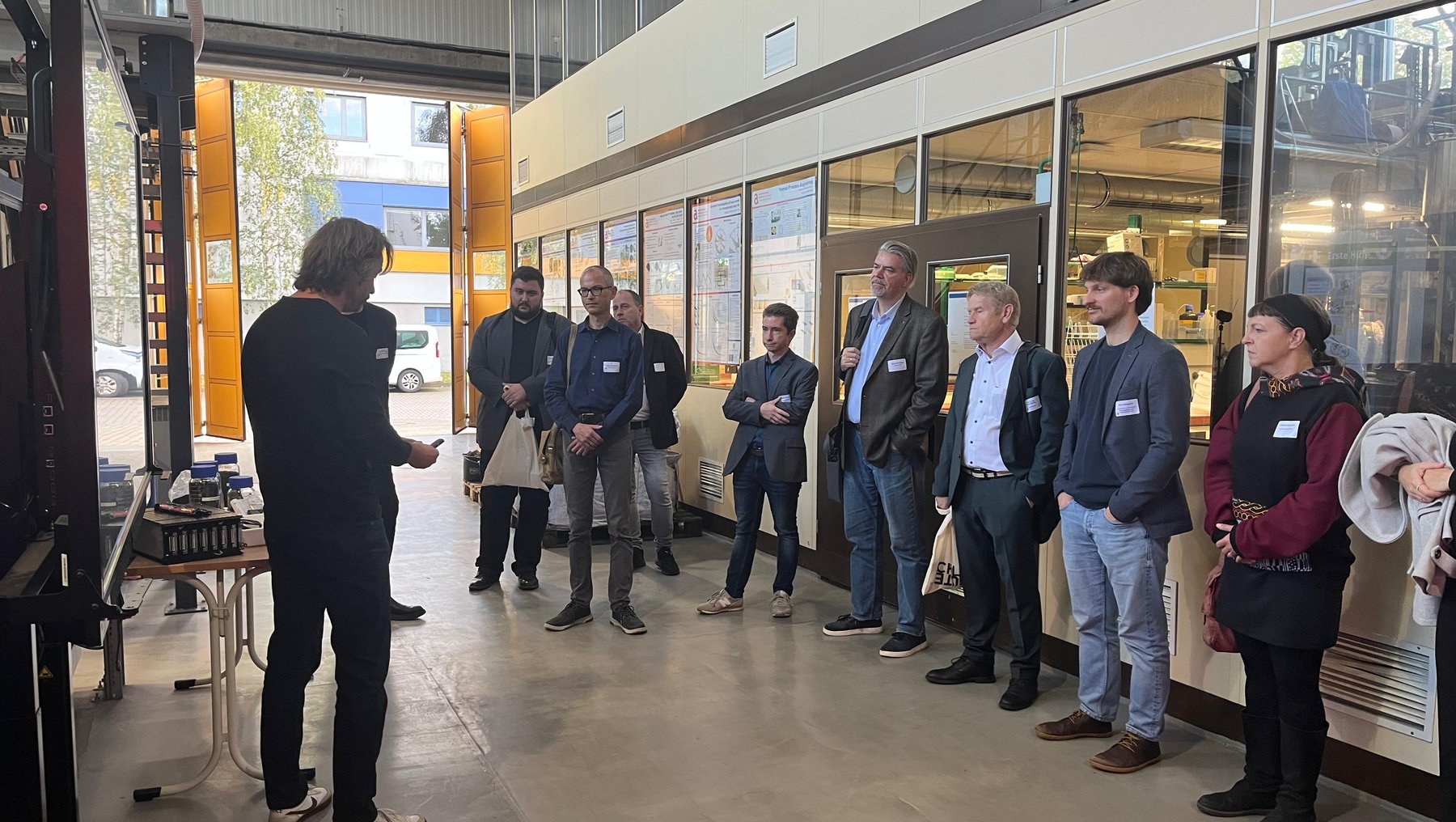 Eine Gruppe von etwa zwölf Personen steht in einer hellen Werkshalle und hört einer Person zu, die vor ihnen steht und etwas erklärt. Im Hintergrund sind Maschinen und Werkbänke zu sehen.