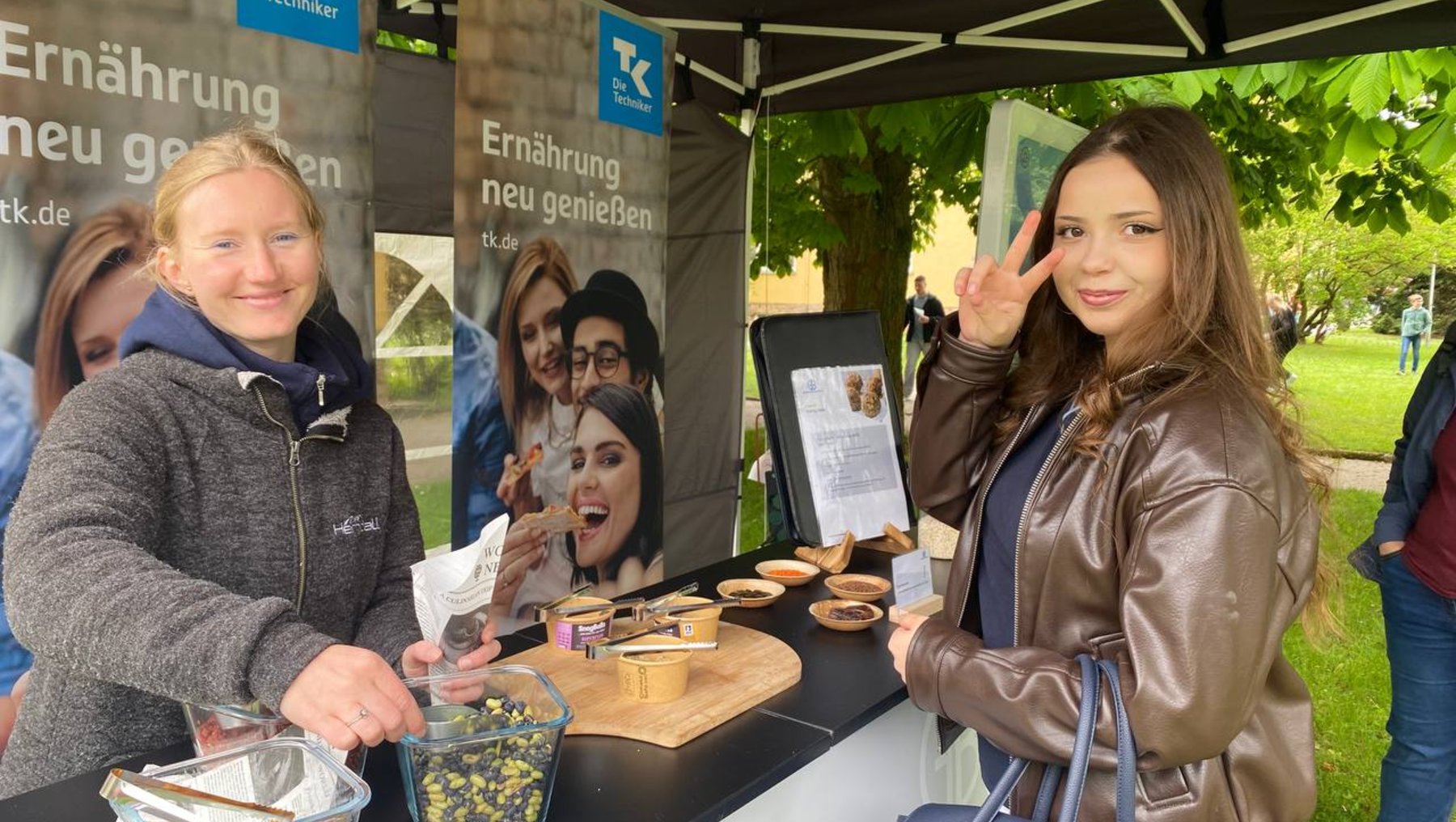 Zwei Frauen stehen an einem Infostand mit dem Thema „Ernährung neu genießen“. Eine probiert Snacks aus einer Schale, die andere macht ein Peace-Zeichen. Der Stand bietet gesunde Lebensmittel und Getränke an.
