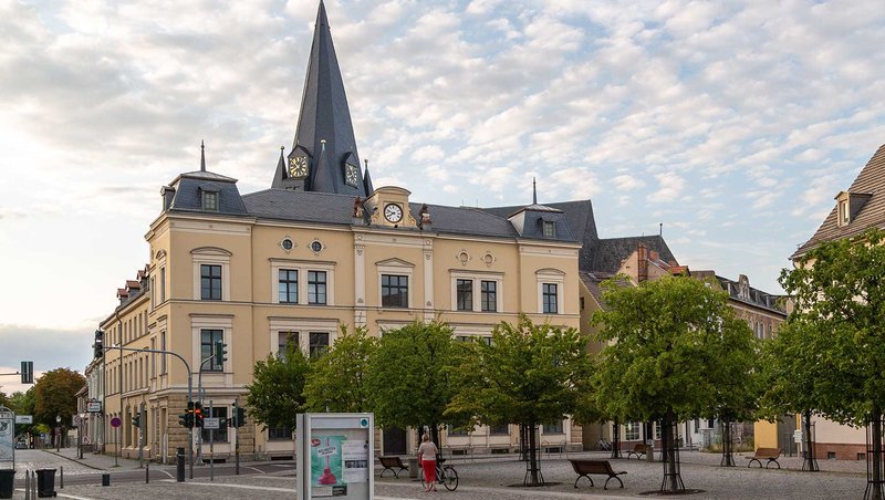 Ein historisches Gebäude mit Uhrturm steht in einer ruhigen Stadtplatzumgebung. Vor dem Gebäude befinden sich Bäume, Bänke und ein Fahrradfahrer. Der Himmel ist leicht bewölkt.