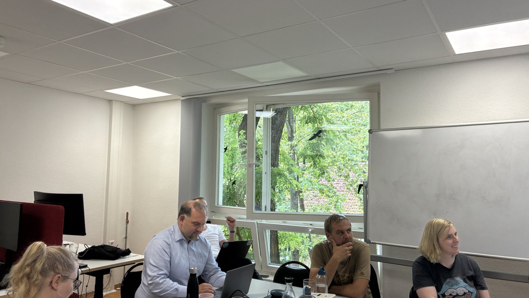Drei Personen sitzen an einem Konferenztisch in einem hellen Raum mit Tageslicht. Vor ihnen liegen Notizblöcke, Stifte und ein Laptop. Eine Person steht mit den Händen in der Hüfte, eine andere schaut nachdenklich, die dritte hält einen Stift. Im Hintergrund ein großer Fensterausschnitt mit Blick auf Bäume.