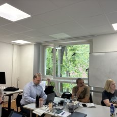 Drei Personen sitzen an einem Konferenztisch in einem hellen Raum mit Tageslicht. Vor ihnen liegen Notizblöcke, Stifte und ein Laptop. Eine Person steht mit den Händen in der Hüfte, eine andere schaut nachdenklich, die dritte hält einen Stift. Im Hintergrund ein großer Fensterausschnitt mit Blick auf Bäume.