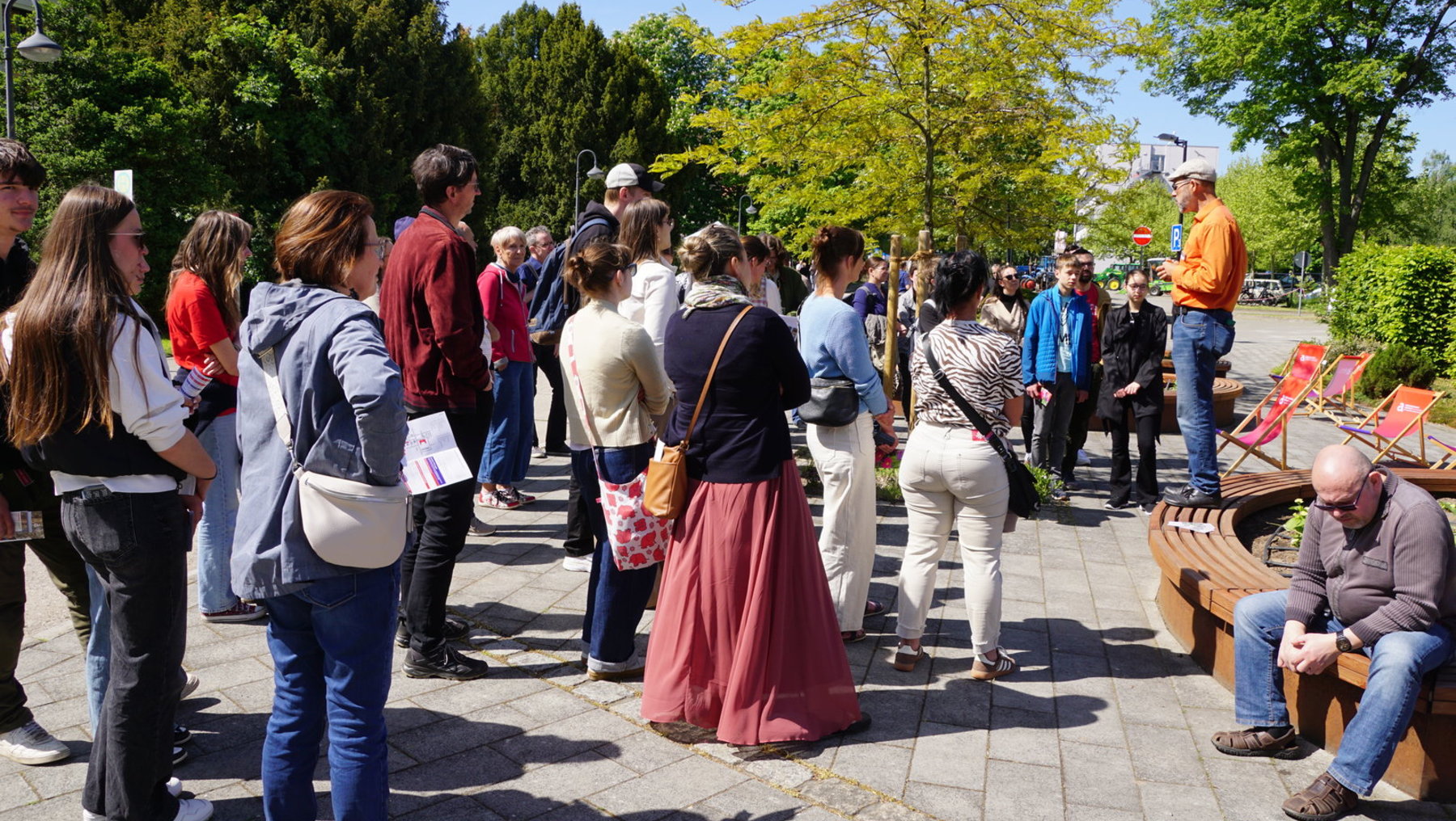 Eine Gruppe von Menschen steht im Freien im Kreis und hört einer Person in historischer Kleidung zu, die vermutlich eine Führung oder einen Vortrag hält. Im Hintergrund sitzt ein älterer Mann auf einer Bank.