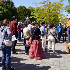 Eine Gruppe von Menschen steht im Freien im Kreis und hört einer Person in historischer Kleidung zu, die vermutlich eine Führung oder einen Vortrag hält. Im Hintergrund sitzt ein älterer Mann auf einer Bank.