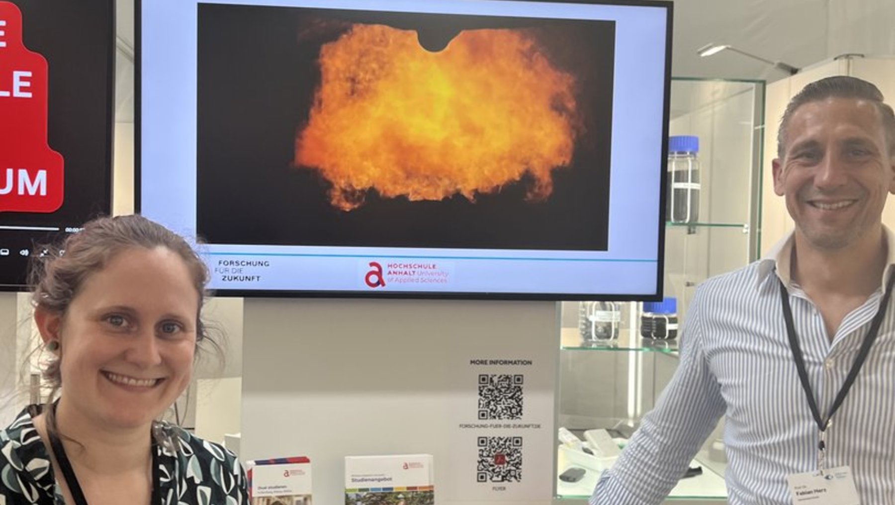 Zwei Personen stehen lächelnd an einem Infostand der Hochschule Anhalt zum Thema „Thermisches Recycling von Lithiumbatterien“. Im Hintergrund ist eine große Flamme auf einem Bildschirm zu sehen, auf dem Tisch liegen Informationsmaterialien und Batterieproben.