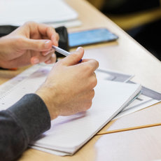 Eine Person sitzt an einem Tisch und hält einen Stift in der Hand, während sie auf einem Blatt Papier schreibt oder markiert. Auf dem Tisch liegen weitere Papiere und ein Smartphone. Die Person trägt einen Pullover mit einem Muster.