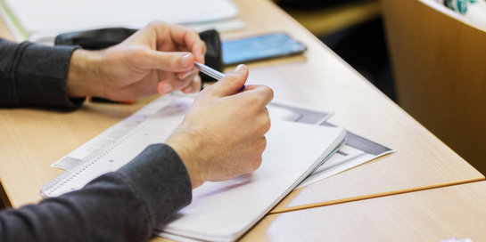 Eine Person sitzt an einem Tisch und hält einen Stift in der Hand, während sie auf einem Blatt Papier schreibt oder markiert. Auf dem Tisch liegen weitere Papiere und ein Smartphone. Die Person trägt einen Pullover mit einem Muster.