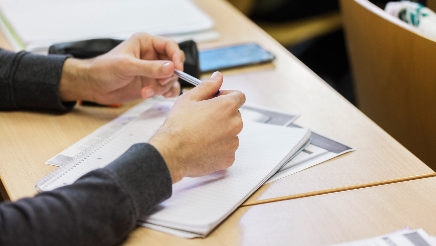 Eine Person sitzt an einem Tisch und hält einen Stift in der Hand, während sie auf einem Blatt Papier schreibt oder markiert. Auf dem Tisch liegen weitere Papiere und ein Smartphone. Die Person trägt einen Pullover mit einem Muster.
