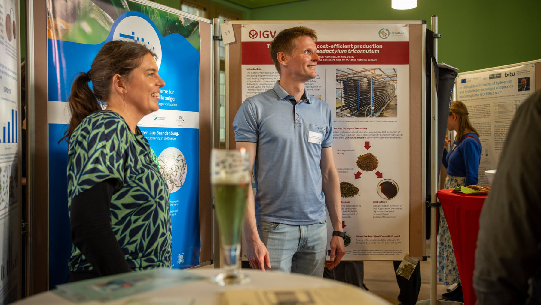 Zwei Personen stehen vor einem Infostand zur kosteneffizienten Produktion von Mikroalgen, insbesondere *Tetraselmis* und *Phaeodactylum tricornutum*. Der Stand zeigt schematisch die Gewinnung von Biomasse, Trocknung und Extraktion von Lipiden für innovative Lebensmittel- und Kosmetikprodukte. Eine Person hält ein Glas mit grünem Algenextrakt.
