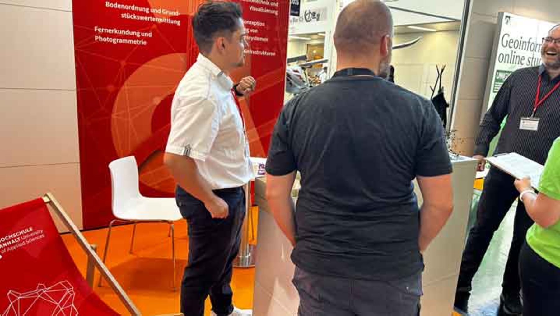 Drei Personen auf einer Messe stehen vor einem Infostand mit rotem Hintergrund und technischen Themen wie Bodenfeuchtemessung und Fernerkundung. Ein Mann im weißen Hemd zeigt etwas auf einem Display, während zwei weitere Personen, einer mit Papier in der Hand, ihm aufmerksam folgen.