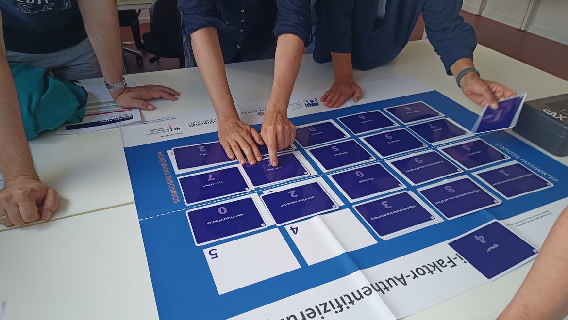 Drei Personen berühren gemeinsam ein großes, blau-weißes Raster mit nummerierten Feldern von 0 bis 5, das als interaktive Faktor-Authentifizierungsvorlage dient. Die Felder sind in einer Matrix angeordnet und werden mit Fingern abgetastet.
