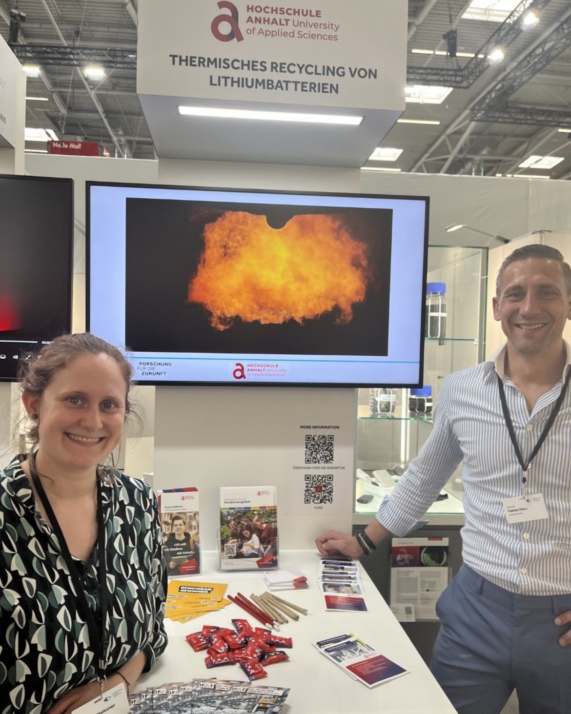 Zwei Personen stehen lächelnd an einem Infostand der Hochschule Anhalt zum Thema Lithium-Batterien recyceln. Im Hintergrund ist eine Explosionsdarstellung auf einem Bildschirm zu sehen, auf dem Tisch liegen Informationsmaterialien und Batterien.
