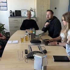 Ein Besprechungstisch mit sieben Personen, die in einem Büroraum sitzen und sich unterhalten. Vor ihnen stehen Getränke, Snacks und Notizblätter, während ein Laptop und ein Telefon auf dem Tisch liegen.