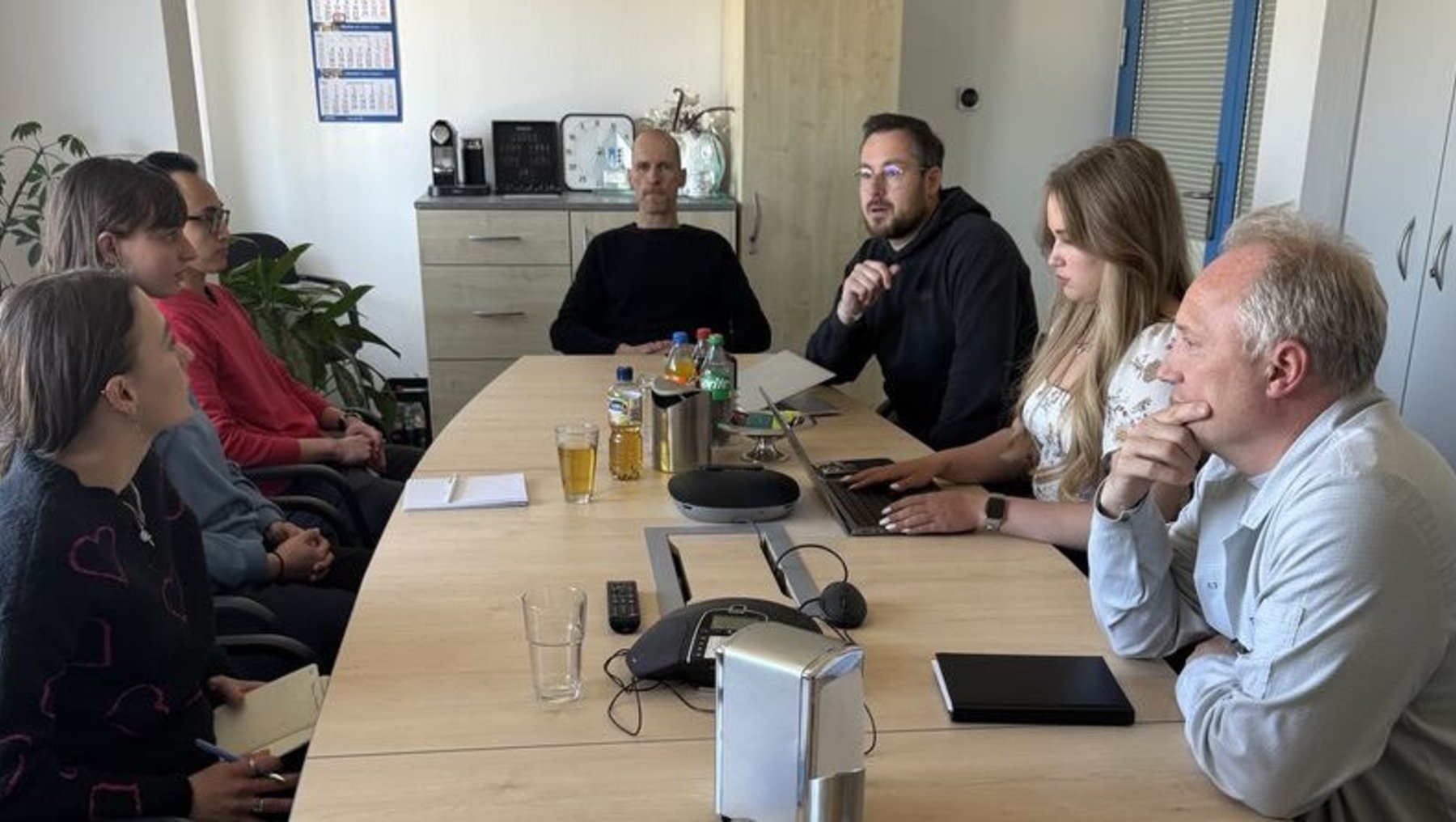 Ein Besprechungstisch mit sieben Personen, die in einem Büroraum sitzen und sich unterhalten. Vor ihnen stehen Getränke, Snacks und Notizblätter, während ein Laptop und ein Telefon auf dem Tisch liegen.